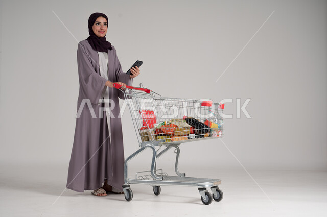 Using a mobile phone while shopping in malls and shopping centers, taking advantage of discounts and sales on products, portrait of a veiled Saudi Arabian Gulf woman wearing a traditional abaya standing and holding a mobile phone in her hand and in front of her is a shopping cart, full body, gray background