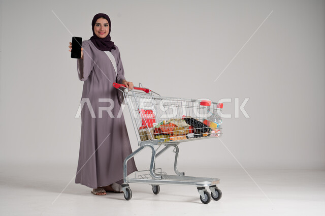 عرض شاشة هاتف محمول سوداء فارغة، الاستمتاع بالتسوق في المولات والمراكز التجارية، الاستفادة من الخصومات والتنزيلات، بورتريه لامرأة عربية خليجية سعودية محجبة ترتدي العباءة التقليدية تقف وتمسك الجوال بيدها وأمامها عربة المشتريات، الجسم بالطول الكامل، خلفية رمادية
