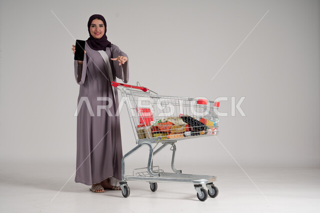 عرض شاشة هاتف محمول سوداء فارغة، الاستمتاع بالتسوق في المولات والمراكز التجارية، الاستفادة من الخصومات والتنزيلات، بورتريه لامرأة عربية خليجية سعودية محجبة ترتدي العباءة التقليدية تقف وتمسك الجوال بيدها وأمامها عربة المشتريات، الجسم بالطول الكامل، خلفية رمادية