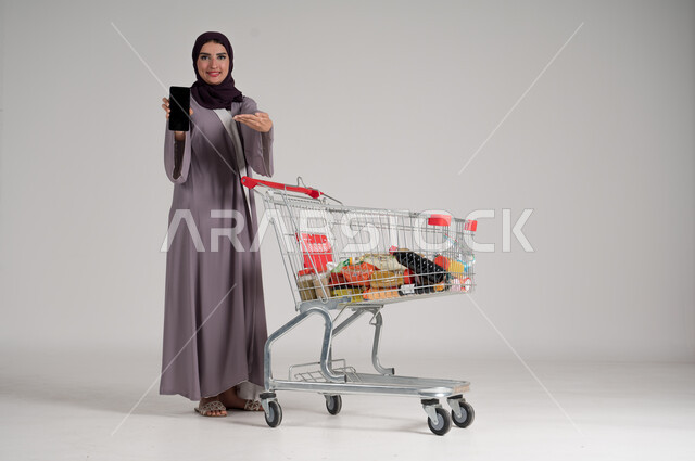 عرض شاشة هاتف محمول سوداء فارغة، الاستمتاع بالتسوق في المولات والمراكز التجارية، الاستفادة من الخصومات والتنزيلات، بورتريه لامرأة عربية خليجية سعودية محجبة ترتدي العباءة التقليدية تقف وتمسك الجوال بيدها وأمامها عربة المشتريات، الجسم بالطول الكامل، خلفية رمادية
