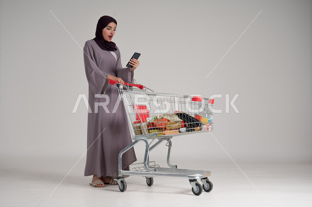 استخدام الهاتف المحمول أثناء التسوق في المولات والمراكز التجارية، الاستفادة من الخصومات والتنزيلات على المنتجات، بورتريه لامرأة عربية خليجية سعودية محجبة ترتدي العباءة التقليدية تقف وتمسك الجوال بيدها وأمامها عربة المشتريات، الجسم بالطول الكامل، خلفية رمادية