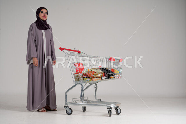 شراء المستلزمات والحاجيات الضرورية، الاستمتاع بالتسوق في المولات والمراكز التجارية، الاستفادة من الخصومات على المنتجات، بورتريه لامرأة عربية خليجية سعودية محجبة ترتدي العباءة التقليدية تقف وأمامها عربة التسوق ببعض المشتريات، الجسم بالطول الكامل، خلفية رمادية