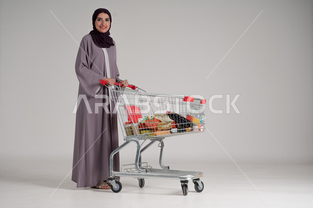 الاستمتاع بالتسوق في المولات والمراكز التجارية، شراء المستلزمات والحاجيات الضرورية، الاستفادة من الخصومات على المنتجات، بورتريه لامرأة عربية خليجية سعودية محجبة ترتدي العباءة التقليدية وتجر عربة التسوق أمامها بإيماءات السعادة، الجسم بالطول الكامل، خلفية رمادية