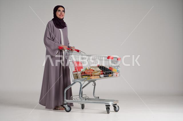 الاستمتاع بالتسوق في المولات والمراكز التجارية، شراء المستلزمات والحاجيات الضرورية، الاستفادة من الخصومات على المنتجات، بورتريه لامرأة عربية خليجية سعودية محجبة ترتدي العباءة التقليدية وتجر عربة التسوق أمامها بإيماءات السعادة، الجسم بالطول الكامل، خلفية رمادية