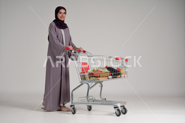 الاستمتاع بالتسوق في المولات والمراكز التجارية، شراء المستلزمات والحاجيات الضرورية، الاستفادة من الخصومات على المنتجات، بورتريه لامرأة عربية خليجية سعودية محجبة ترتدي العباءة التقليدية وتجر عربة التسوق أمامها بإيماءات السعادة، الجسم بالطول الكامل، خلفية رمادية