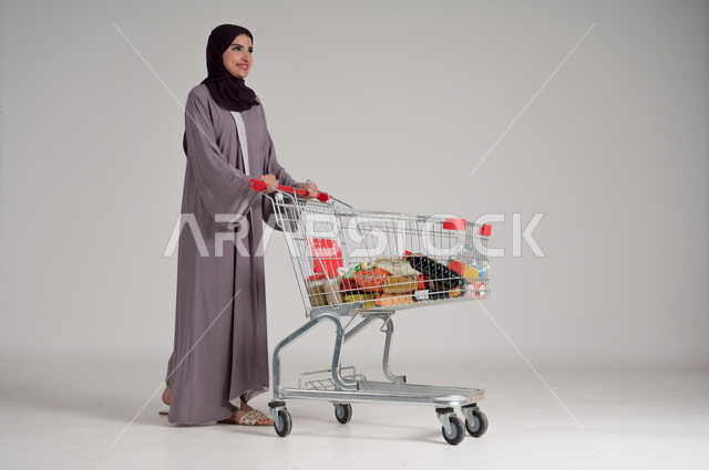 الاستمتاع بالتسوق في المولات والمراكز التجارية، شراء المستلزمات والحاجيات الضرورية، الاستفادة من الخصومات على المنتجات، بورتريه لامرأة عربية خليجية سعودية محجبة ترتدي العباءة التقليدية وتجر عربة التسوق أمامها بإيماءات السعادة، الجسم بالطول الكامل، خلفية رمادية