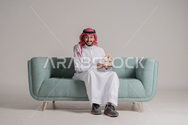 النظر الى الكاميرا بتعابير الفرح، امتلاك سكن حديث والاستقرار العائلي، مفهوم الاسكان والتمليك، ايماءات السعادة بشراء عقار جديد, بورتريه لرجل عربي خليجي سعودي يرتدي الثوب التقليدي والشماغ يجلس على كنبه مريحة ويحمل بيده مجسم بيت جديد، خلفية رمادية
