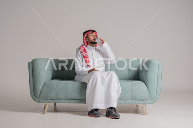 Maintaining and treating kidney health, the importance of drinking pure water to maintain fresh skin, portrait of a Saudi Arabian Gulf man wearing a traditional shemagh and thobe sitting on a comfortable sofa and drinking water from a plastic bottle, maintaining the health of the immune system, getting rid of toxins in the body, gray background