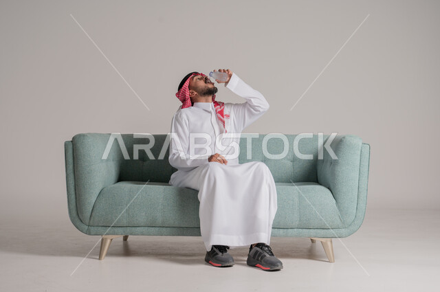 أهمية شرب الماء النقي للمحافظة على بشرة نضرة، بورتريه لرجل عربي خليجي سعودي يرتدي الشماغ والثوب التقليدي يجلس على كنبه مريحه ويشرب المياه من عبوة بلاستيكية، المحافظة على سلامة الجهاز المناعي، التخلص من سموم الجسم، الحفاظ على صحة الكلى ومعالجتها، خلفية رمادية