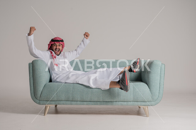 السرور والبشاشة لتحقيق الفوز والانتصار, النظر إلى الكاميرا بتفاعل واندماج وحماس, بورتريه لرجل عربي خليجي سعودي يرتدي الثوب التقليدي والشماغ يستلقي على كنبة مريحة, الشغف بمتابعة المباريات الرياضية, صورة الجسم بالطول الكامل, خلفية رمادية