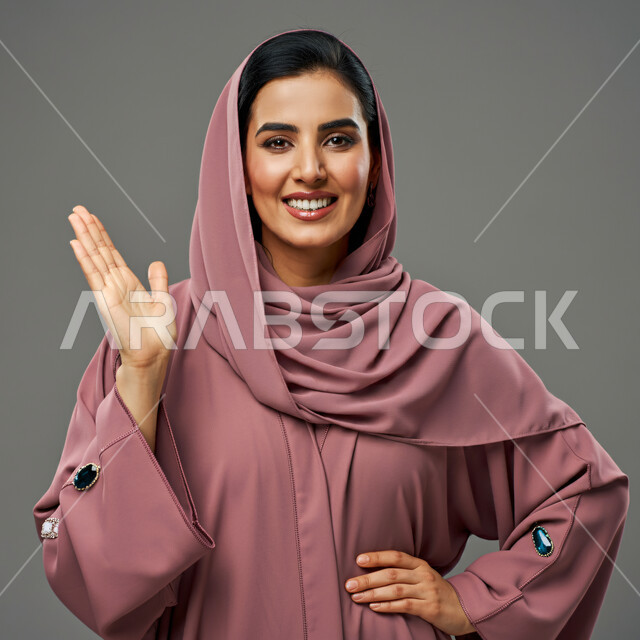 إيماءات الترحيب والسلام، مفهوم الأنوثة والنعومة، الاهتمام بالأناقة والمظهر الخارجي، بورتريه مقرب لإمرأة عربية خليجية سعودية مبتسمة ترتدي الحجاب والعباءة الوردية ترفع يدها للأعلى تنظر الى الكاميرا بتعابير السرور، خلفية رمادية