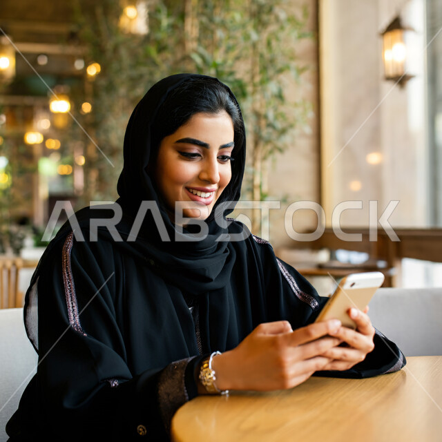 استخدام الأجهزة التقنية الحديثة, قضاء الأوقات الممتعة في المطعم, صورة مقربة لإمرأة عربية خليجية سعودية ترتدي الحجاب والعباءة السوداء تجلس على الطاولة وتستخدم الهاتف المحمول, ايماءات السعادة والسرور