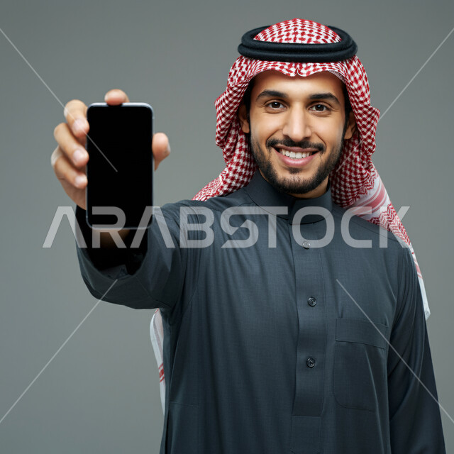 برامج التواصل الاجتماعي عبر الهاتف, عرض تطبيقات و خدمات الهاتف المحمول، استخدام التكنولوجيا المتطورة للتسويق الرقمي الإلكتروني، بورتريه لشاب عربي خليجي سعودي مبتسم يرتدي الشماغ و الثوب التقليدي يحمل بيده الجهاز الجوال بشاشة سوداء فارغة، خلفية رمادية