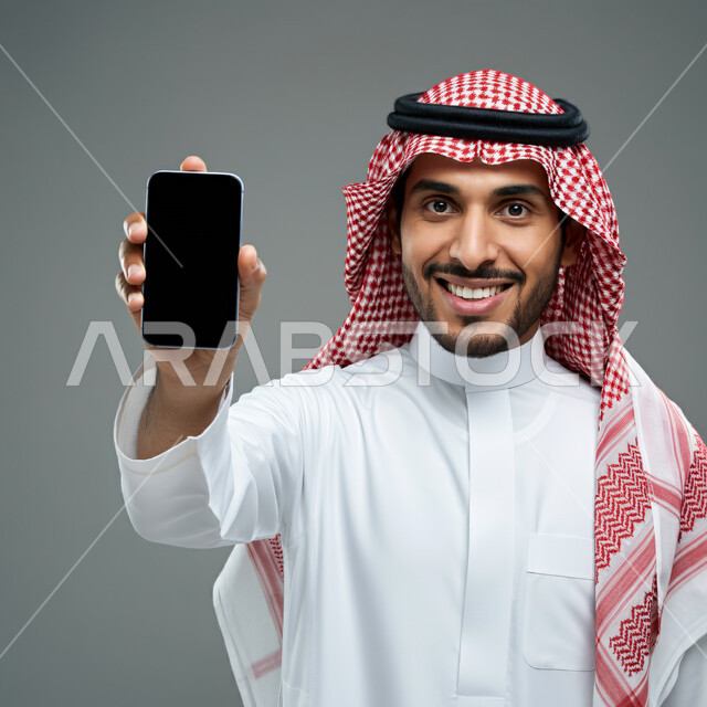 برامج التواصل الاجتماعي عبر الهاتف, عرض تطبيقات و خدمات الهاتف المحمول، استخدام التكنولوجيا المتطورة للتسويق الرقمي الإلكتروني، بورتريه لشاب عربي خليجي سعودي مبتسم يرتدي الشماغ و الثوب التقليدي يحمل بيده الجهاز الجوال بشاشة سوداء فارغة، خلفية رمادية