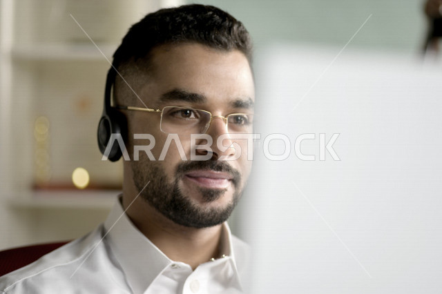 موظف عربي خليجي سعودي يعمل في خدمة العملاء ويرتدي سماعة الاتصال، استعلامات واستفسارات، الاجابه على اسئلة العملاء، اجراء اتصال فيديو من خلال الحاسوب، نظارة طبية، متابعة وتطوير العمل