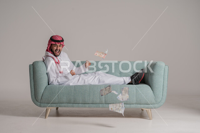 مفهوم الوفرة المالية, التخطيط في تمويل المشاريع المستقبلية, بورتريه لرجل عربي خليجي سعودي مبتسم يرتدي الثوب التقليدي والشماغ يستلقي على كنبة مريحة بإيماءات السرور والسعادة بالإدخار المالي, النمو والأرباح المالية, الاستثمار في الاقتصاد السعودي, خلفية رمادية