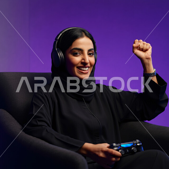 إيماءات وتعابير وجه ويدين تدل على الفوز والانتصار، قضاء أوقات الفراغ في الفيديو غيمز، بورتريه مقرب لإمرأة عربية سعودية خليجية ترتدي الحجاب والعباءة تجلس على كرسي مريح وتحمل بيدها عصا التحكم وتلعب بألعاب الفيديو، خلفية بنفسجية