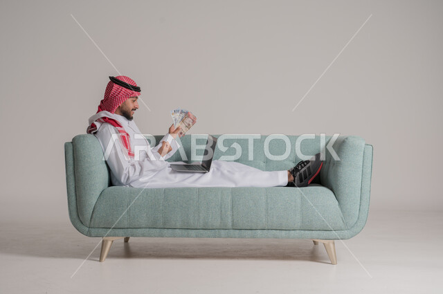 التخطيط في تمويل المشاريع المستقبلية, بورتريه لرجل عربي خليجي سعودي يرتدي الثوب التقليدي والشماغ يستلقي على كنبة يحمل بيديه عملة ورقية للريال السعودي ويعمل على جهاز الحاسب المحمول، إيماءات السرور والسعادة بالإدخار المالي, مفهوم الوفرة المالية, خلفية رمادية