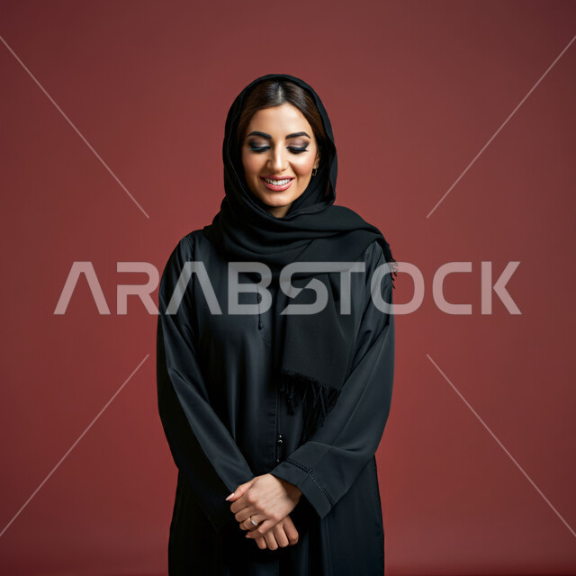 إيماءات السعادة والسرور، الأناقة والاهتمام بالمظهر الخارجي، بورتريه مقرب لامرأة عربية خليجية سعودية ترتدي العباءة السوداء تنظر الى الاسفل بتعابير الخجل والحياء، نظرات الأنوثة الناعمة، خلفية حمراء