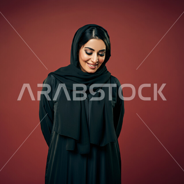 إيماءات السعادة والسرور، الأناقة والاهتمام بالمظهر الخارجي، بورتريه مقرب لامرأة عربية خليجية سعودية ترتدي العباءة السوداء تنظر الى الاسفل بتعابير الخجل والحياء، نظرات الأنوثة الناعمة، خلفية حمراء
