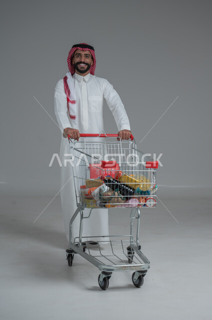 الاستمتاع بالتسوق في المولات والمراكز التجارية، شراء المستلزمات والحاجيات الضرورية، الاستفادة من الخصومات على المنتجات، بورتريه لرجل عربي خليجي سعودي  يرتدي الثوب التقليدي و الشماغ ويجر عربة التسوق أمامه بإيماءات السعادة، الجسم بالطول الكامل، خلفية رمادية