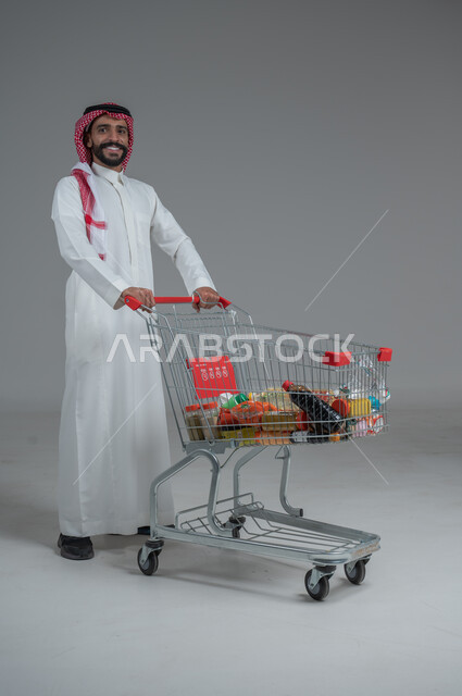 الاستمتاع بالتسوق في المولات والمراكز التجارية، شراء المستلزمات والحاجيات الضرورية، الاستفادة من الخصومات على المنتجات، بورتريه لرجل عربي خليجي سعودي  يرتدي الثوب التقليدي و الشماغ ويجر عربة التسوق أمامه بإيماءات السعادة، الجسم بالطول الكامل، خلفية رمادية