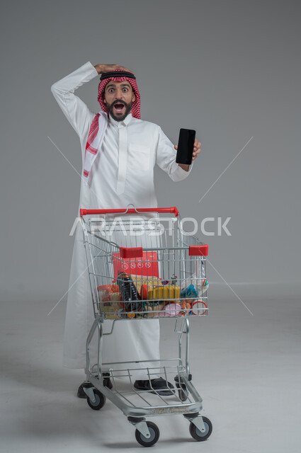 عرض شاشة هاتف محمول سوداء فارغة، الاستمتاع بالتسوق في المولات والمراكز التجارية، الاستفادة من الخصومات والتنزيلات، بورتريه رجل عربي خليجي سعودي يرتدي الثوب التقليدي و الشماغ يقف ويمسك الجوال بيده امام عربة المشتريات بإيماءات الصدمة، الجسم بالطول الكامل، خلفية رمادية