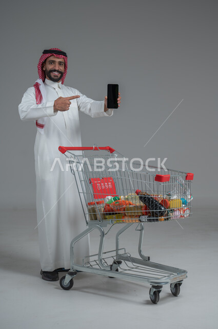  الاستمتاع بالتسوق في المولات والمراكز التجارية، عرض شاشة هاتف محمول سوداء فارغة، الاستفادة من الخصومات والتنزيلات، بورتريه لرجل عربي خليجي سعودي يرتدي الثوب التقليدي و الشماغ يقف ويمسك الجوال بيده امام عربة المشتريات بإيماءات السرور، الجسم بالطول الكامل، خلفية رمادية