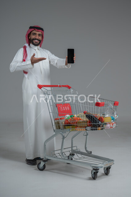  الاستمتاع بالتسوق في المولات والمراكز التجارية، عرض شاشة هاتف محمول سوداء فارغة، الاستفادة من الخصومات والتنزيلات، بورتريه لرجل عربي خليجي سعودي يرتدي الثوب التقليدي و الشماغ يقف ويمسك الجوال بيده امام عربة المشتريات بإيماءات السرور، الجسم بالطول الكامل، خلفية رمادية