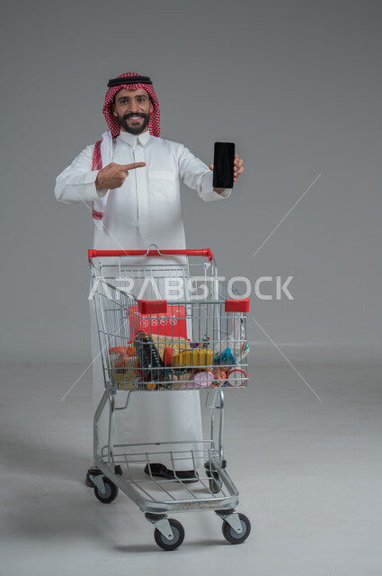  الاستمتاع بالتسوق في المولات والمراكز التجارية، عرض شاشة هاتف محمول سوداء فارغة، الاستفادة من الخصومات والتنزيلات، بورتريه لرجل عربي خليجي سعودي يرتدي الثوب التقليدي و الشماغ يقف ويمسك الجوال بيده امام عربة المشتريات بإيماءات السرور، الجسم بالطول الكامل، خلفية رمادية