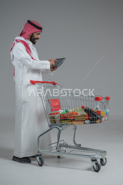 Enjoying shopping in malls and shopping centers, taking advantage of discounts and sales, portrait of a Saudi Arabian Gulf man wearing traditional thobe and shemagh standing and holding a tablet in his hand in front of the shopping cart with gestures of pleasure, full body, gray background