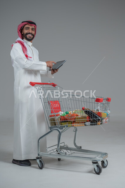 Enjoying shopping in malls and shopping centers, taking advantage of discounts and sales, portrait of a Saudi Arabian Gulf man wearing traditional thobe and shemagh standing and holding a tablet in his hand in front of the shopping cart with gestures of pleasure, full body, gray background