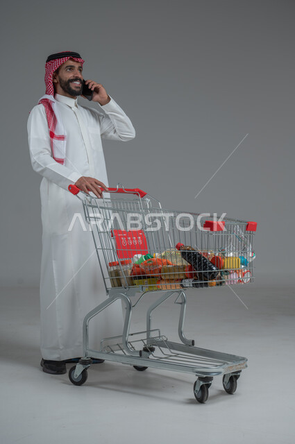  الاستفادة من الخصومات والتنزيلات، الاستمتاع بالتسوق في المولات والمراكز التجارية، بورتريه لرجل عربي خليجي سعودي يرتدي الثوب التقليدي و الشماغ يقف ويمسك الجوال المحمول بيده يجري مكالمة هاتفية بإيماءات السرور و السعادة، الجسم بالطول الكامل، خلفية رمادية