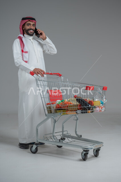 Taking advantage of discounts and sales, enjoying shopping in malls and shopping centers, portrait of a Saudi Arabian Gulf man wearing traditional thobe and shemagh standing and holding a mobile phone in his hand making a phone call with gestures of pleasure and happiness, full body, gray background