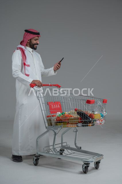  الاستفادة من الخصومات والتنزيلات، الاستمتاع بالتسوق في المولات والمراكز التجارية، بورتريه لرجل عربي خليجي سعودي يرتدي الثوب التقليدي و الشماغ يقف ويمسك الجوال المحمول بيده بإيماءات السرور و السعادة، الجسم بالطول الكامل، خلفية رمادية