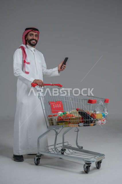 Taking advantage of discounts and sales, enjoying shopping in malls and shopping centers, portrait of a Saudi Arabian Gulf man wearing traditional thobe and shemagh standing and holding a mobile phone in his hand with gestures of pleasure and happiness, full body, gray background
