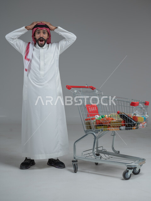  الاستمتاع بالتسوق في المولات والمراكز التجارية، الاستفادة من الخصومات والتنزيلات، بورتريه لرجل عربي خليجي سعودي يرتدي الثوب التقليدي و الشماغ يقف ويضع يديه على رأسه امام عربة المشتريات بإيماءات الصدمة والدهشة، الجسم بالطول الكامل، خلفية رمادية