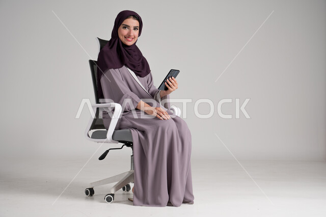 Looking at camera with gestures of pleasure, using modern devices and technologies in daily life, portrait of a veiled Saudi Arabian Gulf woman wearing traditional abaya sitting on a swivel chair holding a mobile phone in her hand, browsing websites, full body, gray background