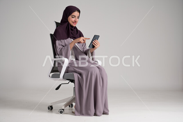 الإشارة إلى الجوال بإيماءات السرور، استخدام الأجهزة والتقنيات الحديثة في الحياة اليومية، بورتريه لامرأة عربية خليجية سعودية محجبة ترتدي العباءة التقليدية تجلس على كرسي دوار تمسك الهاتف المحمول بيدها و تتصفح مواقع الانترنت، الجسم بالطول الكامل، خلفية رمادية