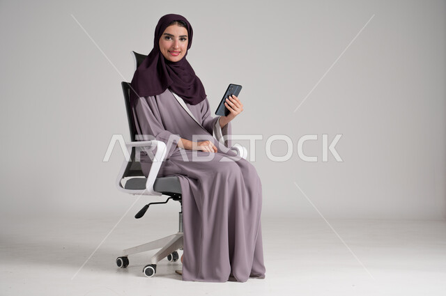 Looking at camera with gestures of pleasure, using modern devices and technologies in daily life, portrait of a veiled Saudi Arabian Gulf woman wearing traditional abaya sitting on a swivel chair holding a mobile phone in her hand, browsing websites, full body, gray background