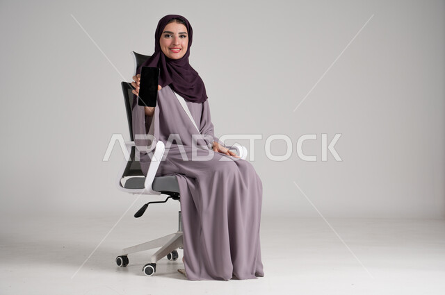 Empty black mobile screen, use of modern devices and technologies in daily life, portrait of a veiled Saudi Arabian Gulf woman wearing traditional abaya sitting on a swivel chair holding a mobile phone in her hand and looking at the camera with cheerful gestures, full body, gray background
