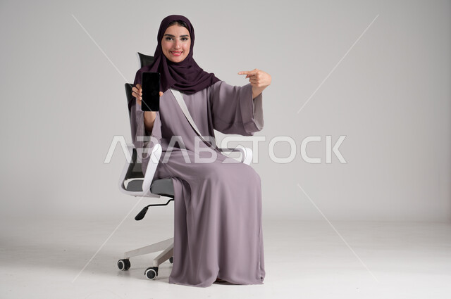 عرض شاشة جوال سوداء فارغة، استخدام الأجهزة والتقنيات الحديثة في الحياة اليومية، بورتريه لامرأة عربية خليجية سعودية محجبة ترتدي العباءة التقليدية تجلس على كرسي دوار تمسك الهاتف المحمول وتشير إليه بيدها بإيماءات البشاشة والثقة، الجسم بالطول الكامل، خلفية رمادية