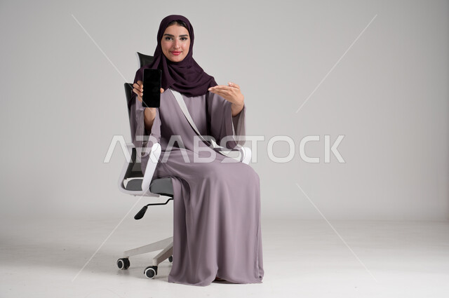 عرض شاشة جوال سوداء فارغة، استخدام الأجهزة والتقنيات الحديثة في الحياة اليومية، بورتريه لامرأة عربية خليجية سعودية محجبة ترتدي العباءة التقليدية تجلس على كرسي دوار تمسك الهاتف المحمول وتشير إليه بيدها بإيماءات البشاشة والثقة، الجسم بالطول الكامل، خلفية رمادية