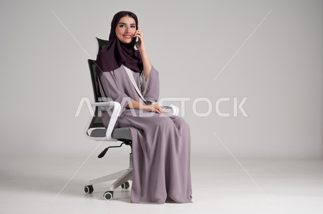 استخدام الأجهزة والتقنيات الحديثة في الحياة اليومية، بورتريه لامرأة عربية خليجية سعودية محجبة ترتدي العباءة التقليدية تجلس على كرسي دوار تجري مكالمة هاتفية عبر الهاتف المحمول، التواصل مع الأهل والأصدقاء وسماع آخر الأخبار، الجسم بالطول الكامل، خلفية رمادية