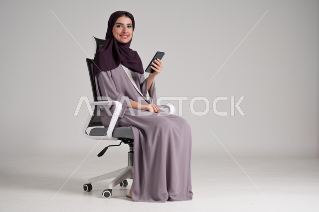 Looking at camera with gestures of pleasure, using modern devices and technologies in daily life, portrait of a veiled Saudi Arabian Gulf woman wearing traditional abaya sitting on a swivel chair holding a mobile phone in her hand, browsing websites, full body, gray background