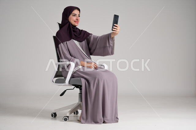 استخدام الأجهزة والتقنيات الحديثة في الحياة اليومية، بورتريه لامرأة عربية خليجية سعودية محجبة ترتدي العباءة التقليدية تجلس على كرسي دوار تجري مكالمة فيديو عبر الهاتف المحمول، التواصل مع الأهل والأصدقاء وسماع آخر الأخبار، الجسم بالطول الكامل، خلفية رمادية