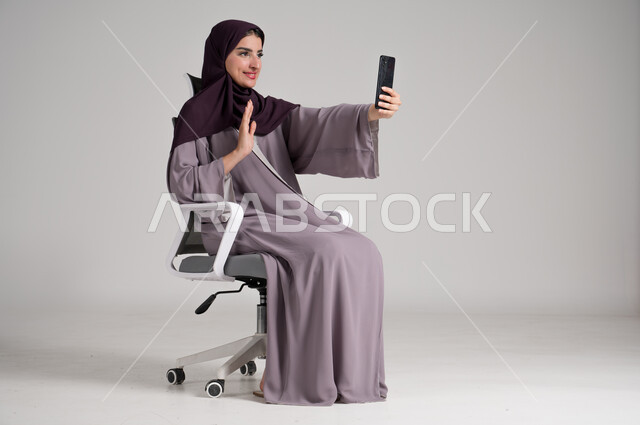 استخدام الأجهزة والتقنيات الحديثة في الحياة اليومية، بورتريه لامرأة عربية خليجية سعودية محجبة ترتدي العباءة التقليدية تجلس على كرسي دوار تجري مكالمة فيديو عبر الهاتف المحمول، التواصل مع الأهل والأصدقاء وسماع آخر الأخبار، الجسم بالطول الكامل، خلفية رمادية