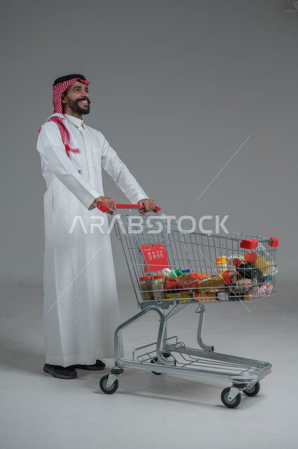 الاستمتاع بالتسوق في المولات والمراكز التجارية، شراء المستلزمات والحاجيات الضرورية، الاستفادة من الخصومات على المنتجات، بورتريه لرجل عربي خليجي سعودي يرتدي الثوب التقليدي و الشماغ ويجر عربة التسوق أمامه بإيماءات السعادة، الجسم بالطول الكامل، خلفية رمادية