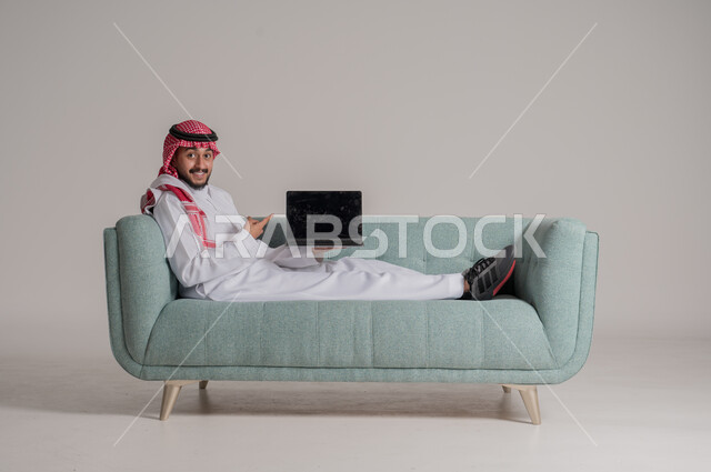 عرض شاشة سوداء فارغة على جهازاللابتوب، النظر الى الكاميرا والاشارة الى جهاز الحاسب المحمول بتعابير وايماءات الاعجاب، بورتريه لرجل عربي خليجي سعودي مبتسم يرتدي الثوب التقليدي والشماغ يستقلي على كنبة ويقوم باستخدام التقنيات الحديثه المتطورة، خلفية رمادية