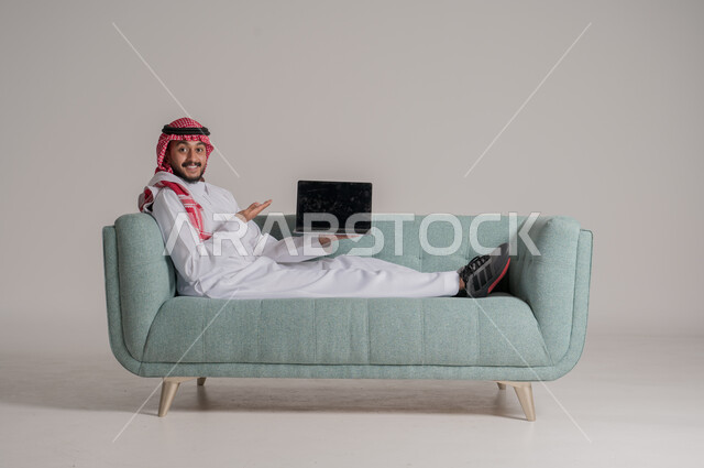 النظر الى الكاميرا والاشارة الى جهاز الحاسب المحمول بتعابير وايماءات الاعجاب، بورتريه لرجل عربي خليجي سعودي مبتسم يرتدي الثوب التقليدي والشماغ يستقلي على كنبة ويقوم باستخدام التقنيات الحديثه المتطورة، عرش شاشة سوداء فارغة على جهازاللابتوب، خلفية رمادية